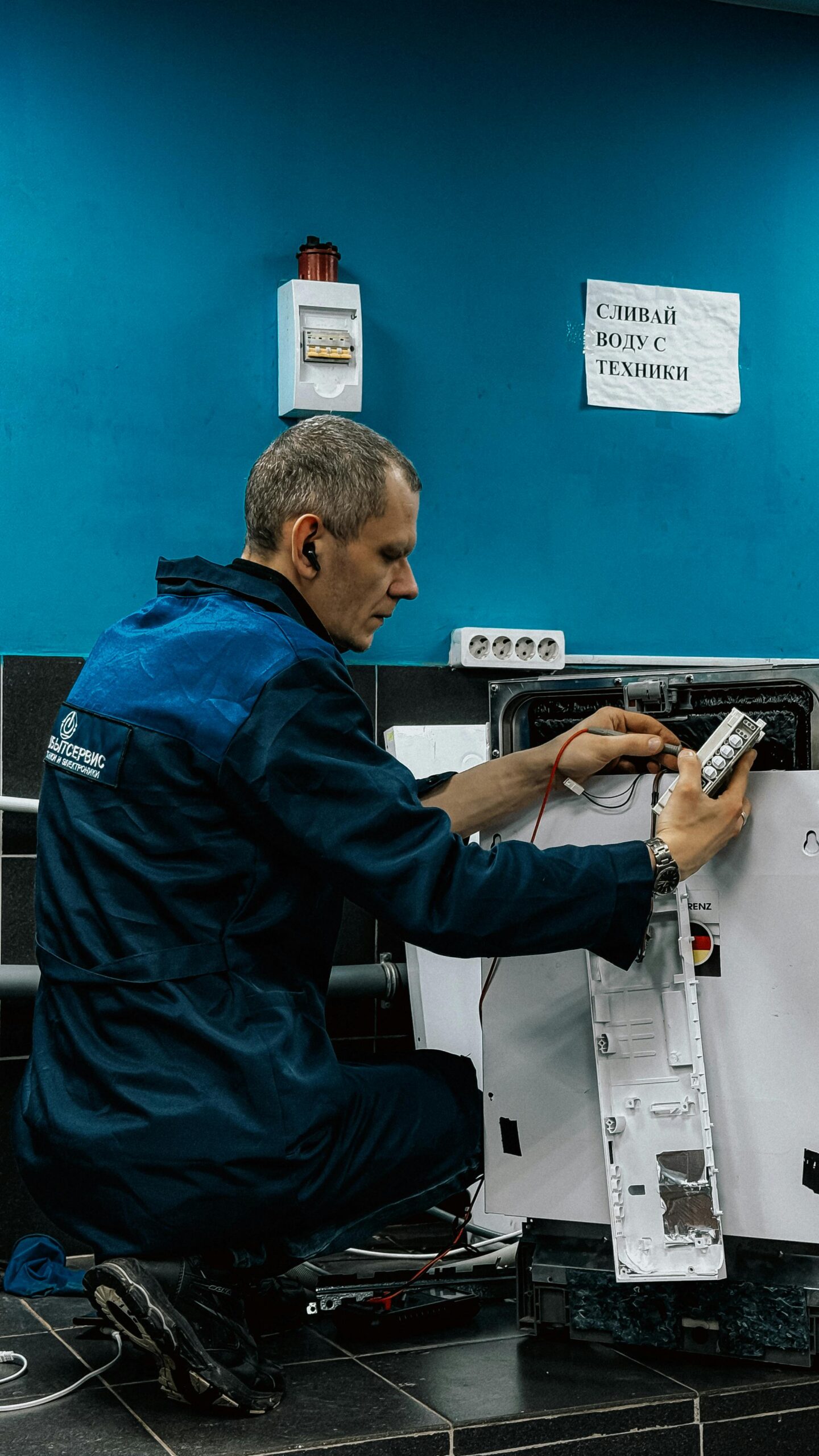 A focused technician in blue uniform repairs a home appliance in an indoor setting.