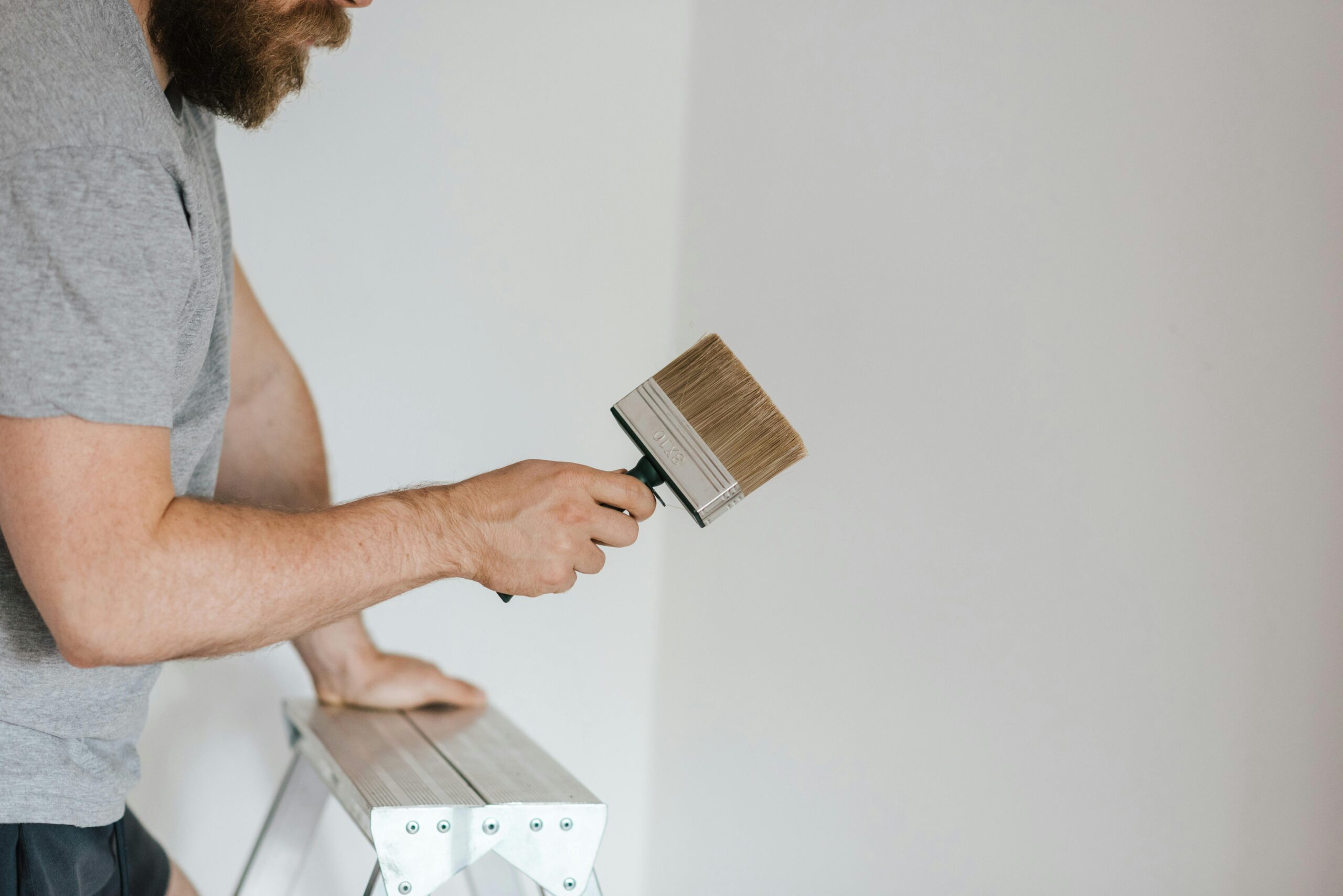 Side view of focused skilled decorator observing brush while leaning on ladder on gray background at daytime